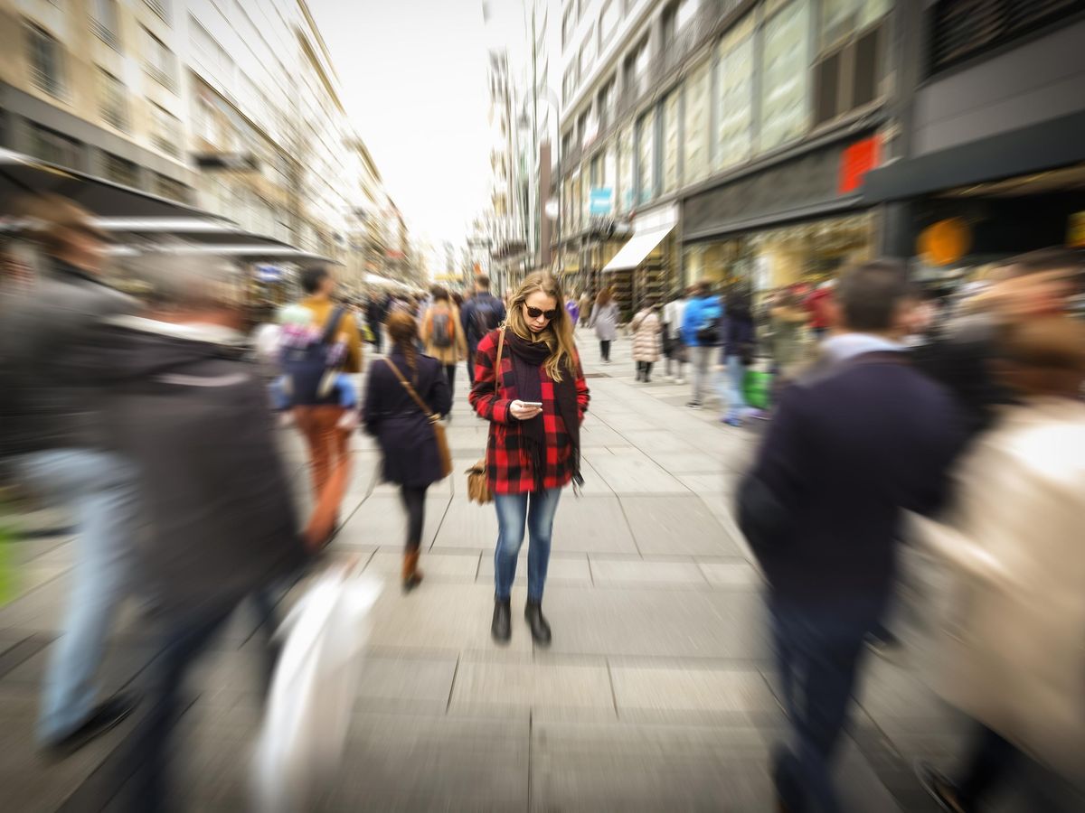 一个穿着红黑外套和牛仔裤的金发女人看着她的手机。她周围的人群和街道模糊了。