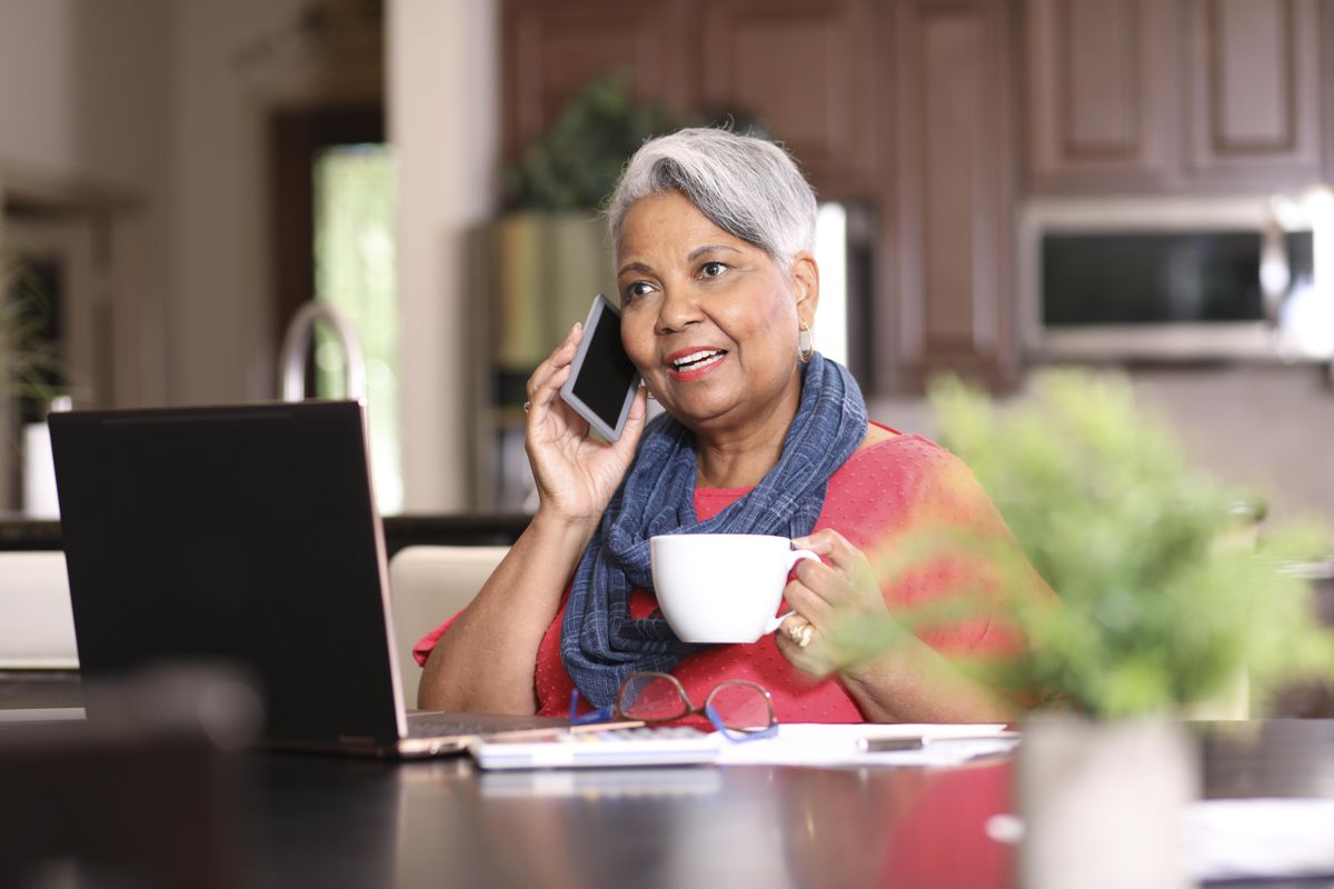 一位老妇人在家里讲电话,在电脑前,手里拿着一杯咖啡